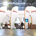 Jamies Exhibition Booth Featuring White Curved Structure Orange Branding Plants And Staff