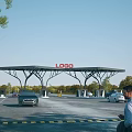 Charging Station Entrance Gate With LOGO And Vehicles Entering Under Blue Sky