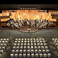 Orchestra Performance On Stage With Wooden Backdrop Bright Lighting And Arranged Seats