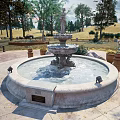Outdoor Stone Garden Fountain With Central Sculpture And Surrounding Flower Pots On Brick Patio Near Trees