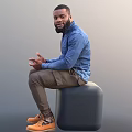 Man In Blue Denim Shirt Brown Pants Sitting On Gray Cube Gesturing With Hands