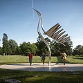 Modern Stainless Steel Landscape Sculpture Resembling Crane in Park Grassland with Trees and People Walking