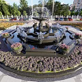 Ornate Circular Fountain With Purple Flower Beds Paved Surroundings And Greenery