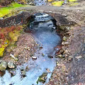 Ancient Stone Bridge Over Flowing Stream With Mossy Rocks And Natural Surroundings
