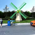 Green Windmill With Yellow Center By Roadside Recycling Bin Orange Sports Car And Trees