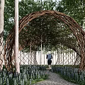 Arch Shaped Woven Branch Pergola with Trees Purple Flowers and People Running on Path