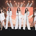 Five Women In White Outfits Singing On Stage With Microphones And Stage Lights