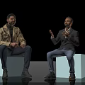 Two men in trench coat and blazer sitting on white cubes gesturing against black background