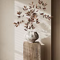 Gray Vase With Brown Leaves On Wooden Cube Base Against Light Wall With Sunlight Shadows