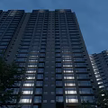 Modern Tall High Rise Apartment Building With Evening Window Lights And Green Trees