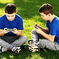 Two Boys Sitting On Grass Playing With Phones Wearing Blue T Shirts And Gray Pants