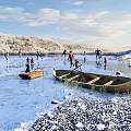 People Ice Skating On Frozen Lake With Boats Near Snowy Hills And Blue Sky