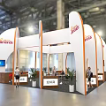 Jamies Exhibition Booth Featuring White Curved Structure Orange Branding Plants And Staff