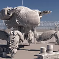 Camouflage Painted Mechanical Crab Sculpture With Cannons Barbed Wire Wooden Crates Oil Drums Blue Sky