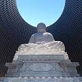 White Buddha Sculpture Seated On Lotus Pedestal Against Curved Black Building Blue Sky