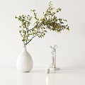 White Vase With Green Foliage Next To White Statue And Small Candle On White Table