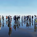 Group of People Standing on Beach with Water Reflections Under Clear Blue Sky
