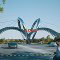 Modern Entrance Gate with Curved Arch Structure Featuring Logo Surrounded by Cars Pedestrians and Trees