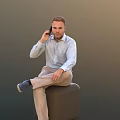 Man Talking On Phone Sitting On Black Cube With Light Blue Shirt And Beige Pants