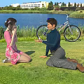 Man And Woman Interact On Grass Lawn By Lake With Bicycle