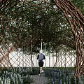 Arch Shaped Woven Branch Pergola with Trees Purple Flowers and People Running on Path