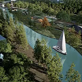 Scenic Waterfront Landscape With River Sailboats Lush Trees And Modern Buildings