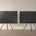 Two Black Modern TVs with Tripod Stands on Wooden Floor Against Neutral Wall