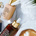 Elegant Cosmetics Display Featuring Lipstick Foundation Facial Serum Gold Necklace Rings White Flowers And Coffee