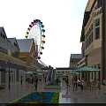 Illuminated Commercial Street With Ferris Wheel Outdoor Seating Pedestrians Flower Beds And Buildings