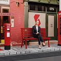 Woman Sitting On Red Bench Near Red Telephone Booth And Postbox By Brick Building