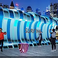 Surfing Themed Illuminated Wave Structure With People Taking Photos In Urban Night View