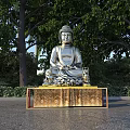 Silver Buddha Sculpture on Golden Pedestal in Outdoor Garden with Trees and Greenery