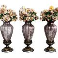 Three Metallic Vases With Decorative Bases Holding White And Beige Flowers With Green Leaves