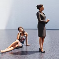 Two Women In Spacious Room One Sitting On Dark Floor And One Standing In Suit