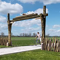 Rustic Wooden Entrance Gate with Log Arch Cyclist Riding Through Countryside Field Under Blue Sky