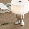 Table Lamp With Silver Humanoid Leg Base White Pleated Lampshade Near Open Book Wooden Floor