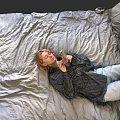 Woman Lying on Gray Bed Sheets Wearing Striped Shirt and Light Blue Jeans Holding Phone