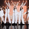 Five Women In White Outfits Singing On Stage With Microphones And Stage Lights