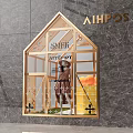 House Shaped Shop Window with Golden Frame featuring Mannequin in Brown Coat and Yellow Bucket