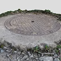 Circular Metal Manhole Cover On Stone Base With Surrounding Weeds And Small Stones