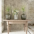 Glass Vases With White Daisies And Green Tiny Flowers On Wooden Table Against Weathered Wall