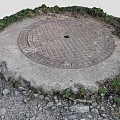 Circular Metal Manhole Cover On Stone Base With Surrounding Weeds And Small Stones