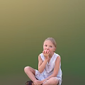 Little Girl Eating Apple In White Sleeveless Dress Seated On Ground Against Green Background