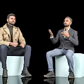 Two men in trench coat and blazer sitting on white cubes gesturing against black background