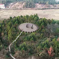 Aerial View Of Circular Building Surrounded By Lush Trees Path Farmland And Distant Houses