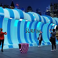 Surfing Themed Illuminated Wave Structure With People Taking Photos In Urban Night View