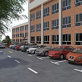 Parking Lot With Multiple Cars Near Red Brick Building And Green Trees