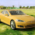Yellow Tesla Sedan Parked On Green Grass Field With Trees And Blue Sky Background
