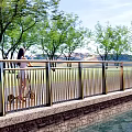 Metal Railing On Waterfront Path With Trees Grassland Mountains And Person Riding Electric Scooter