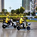 Men Wearing Yellow Uniforms Riding Electric Scooters in City Street with High Rise Buildings
